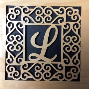 May include: A wooden plaque with a decorative cutout design featuring a large letter L in the center. The letter is cut out of the wood and the background is a black material.