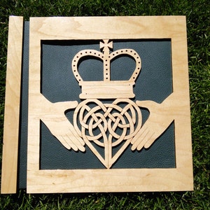 May include: A wooden plaque featuring a carved design of two hands clasped around a heart with a crown on top. The background is a dark blue fabric.