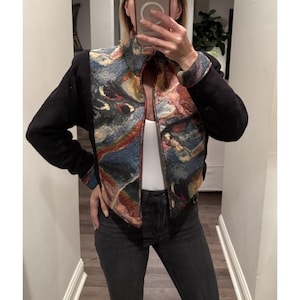 May include: A black jacket with a colorful tapestry-like design on the front and collar. The jacket has long black sleeves and a high collar. The colorful design includes shades of blue, red, and brown. The jacket is zipped up.