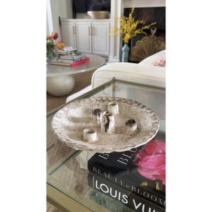 May include: A round, off-white ceramic candle holder with a textured, speckled finish and a scalloped edge. The piece features five small, cylindrical candle sockets arranged in the center, each with dark, vertical streaks of brown and black.