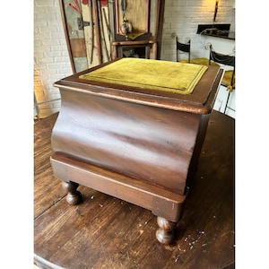 May include: A vintage wooden footstool with a dark brown finish. The top is upholstered in a gold-colored velvet fabric. The footstool has a curved design on the sides and rests on four small, rounded feet.