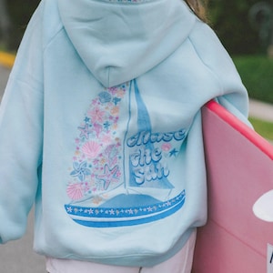 Könnte beinhalten: Hellblauer Kapuzenpullover mit Segelboot-Design auf dem Rücken, mit dem Text "Chase the Sail". Das Segelboot ist mit Muscheln und Seesternen in Rosa und Blau verziert. Die Kapuze hat ein Sternmuster.