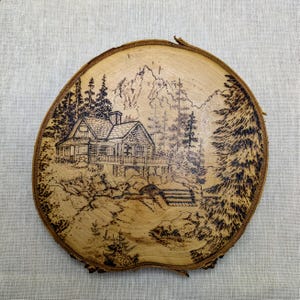Op de afbeelding: Een ronde houten schijf met een pyrografie landschap. Het kunstwerk toont een hut omringd door bomen, met bergen op de achtergrond. Het stuk heeft een natuurlijke schorsrand en een warme, natuurlijke houttint.