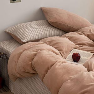 May include: A bed with a blush-colored comforter and pillows. A striped pillow and bed skirt are also visible. A red apple rests on an open book on the bed, creating a cozy and inviting scene.