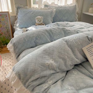 May include: A light blue duvet cover and pillow set with a bow pattern. The set includes a white ruffled edge. A small teddy bear sits on the bed. The bedding has a textured appearance. The duvet and pillows are in a bedroom setting.