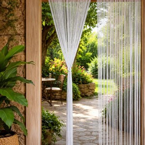 May include: A white string curtain made of numerous thin, vertical strands hanging from a wooden door frame. The curtain is gathered and tied together at the center, creating a draped effect that allows a view of an outdoor garden area.