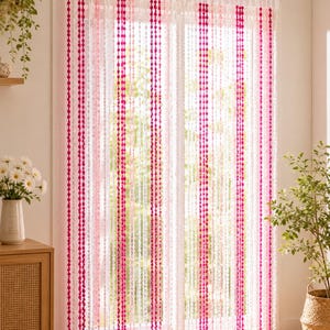 Peut inclure: Un rideau de fenêtre transparent composé de brins verticaux de pompons dans des tons alternés de rose vif, rose clair et blanc. Le rideau est suspendu à une tringle argentée et se compose de multiples cordons suspendus formant un motif rayé.