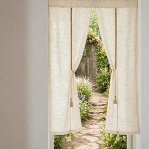 May include: Beige linen curtains framing a view of a garden path. The curtains are tied back with rope, revealing a stone path, wooden fence, and lush greenery. The curtains have a simple, natural design.