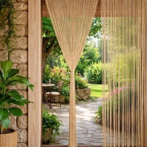 Può includere: Tenda a filo beige appesa in una porta, con vista su un giardino. La tenda è composta da molti fili verticali. La porta è incorniciata da un bordo in legno e un muro di pietra. È visibile un rigoglioso giardino con fiori e vegetazione.