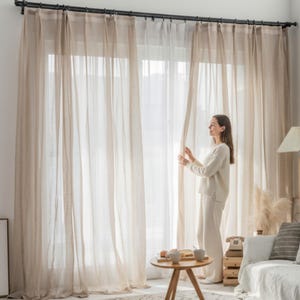 Op de afbeelding: Beige transparante gordijnen die aan een donkere metalen stang hangen. De gordijnen zijn lang en vloeiend, waardoor zacht, diffuus licht ontstaat. Een persoon staat bij de gordijnen en houdt ze open. De kamer heeft een neutraal kleurenpalet met een houten tafel en een lamp.