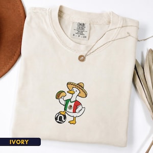 May include: An ivory t-shirt featuring an embroidered graphic of a white duck wearing a sombrero and a shirt with the colors of the Mexican flag. The duck holds a taco in one wing and rests a foot on a black and white soccer ball. The shirt tag reads Comfort Colors.