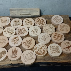 Könnte beinhalten: Eine Sammlung von Holzuntersetzern mit eingravierten Designs und Texten. Die Untersetzer zeigen verschiedene Phrasen und Bilder, darunter Campingszenen, Grillwerkzeuge und humorvolle Sprüche. Die Untersetzer sind auf einer Holzoberfläche angeordnet.