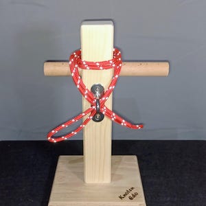 Könnte beinhalten: Ein hölzernes Knotenbinde-Display mit einem rot-weißen Seil, das um einen Metallring gebunden ist. Das Display hat einen Holzsockel und einen vertikalen Pfosten mit einem horizontalen Dübel. Der Text "Knoten Edo" ist auf dem Sockel sichtbar.