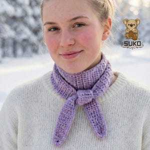 May include: A light purple crocheted scarf tied in a knot. The scarf is made of a textured yarn and is worn over a cream-colored sweater. The image includes the SUKO logo with a koala bear knitting.