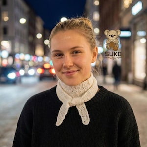 Op de afbeelding: Een vrouw draagt een witte, handgebreide sjaal die in een knoop is gelegd. De sjaal is gemaakt van een lichtgekleurd garen en wordt over een zwarte trui gedragen. De achtergrond is een vage stadsstraat 's nachts.