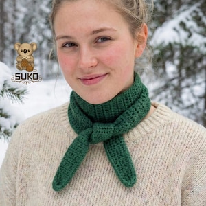 May include: A green crochet scarf tied in a knot, worn over a cream-colored sweater. The scarf is a deep shade of green and appears to be handmade. The background shows a snowy, outdoor scene. The logo "SUKO" is visible in the upper left corner.