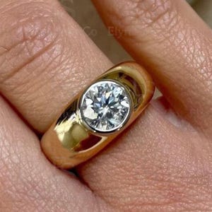 Può includere: Un anello d'oro con una grande gemma rotonda e trasparente incastonata in una lunetta. L'anello ha una fascia spessa e arrotondata e una finitura lucida. La gemma brilla, riflettendo la luce.