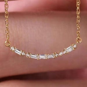以下が含まれることがあります： 小さな透明なバゲットカットの石が並んだ、カーブしたバーペンダントが付いた繊細なゴールドチェーンネックレス。ネックレスは細いリンクチェーンのデザインで、ペンダントは緩やかな弧を描いており、控えめで洗練された外観を作り出しています。