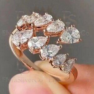 May include: Rose gold ring with multiple pear-shaped clear gemstones. The gemstones are arranged in a flowing, asymmetrical design. The ring is held by a hand against a grey background. The ring's design is elegant and eye-catching.