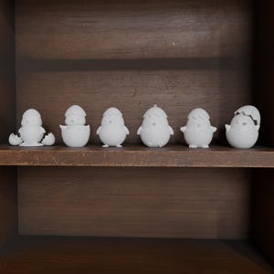 May include: A collection of seven white ceramic Easter chick figurines in various stages of hatching, displayed on a dark wooden shelf. The chicks are in different poses, some emerging from cracked eggs, others standing upright.