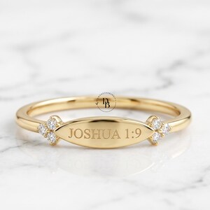 Pode incluir: Um anel de ouro com uma placa oval horizontal central gravada com o texto JOSHUA 1:9. A banda é decorada em ambos os lados da placa por um grupo de três pequenas pedras claras de corte redondo em uma formação triangular.