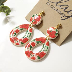 Op de afbeelding: Een paar bloemen oorbellen. De oorbellen hebben een witte basis met een rood bloemenpatroon en groene bladeren. De oorbellen hebben een goudkleurige pin en hebben de vorm van een cirkel en een ovaal.