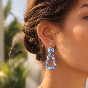 May include: A pair of dangle earrings featuring a small circular stud and a larger trapezoidal drop piece. Both components are made of white material with a blue floral pattern and are connected by a small gold-toned metal jump ring.