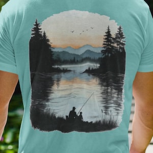 May include: A light blue t-shirt featuring a scenic illustration of a lake at sunset. The design shows silhouettes of trees, mountains, and two people fishing. The shirt's design is a nature-themed graphic.