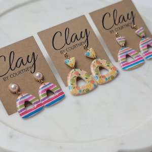 May include: Handmade clay earrings in various designs. One pair features a pearl stud and a rainbow striped arch. Another has a floral pattern with a triangle top and a teardrop shape. The earrings are displayed on brown cards with the text "Clay BY COURTNEY".