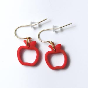 May include: A pair of drop earrings featuring bright red, hollow apple-shaped charms. Each charm is suspended from a gold-toned metal hook wire, which includes a small, clear silicone backing to secure the earring in place during wear.