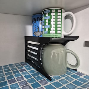 May include: A black plastic shelf organiser with a slatted side design. On the top shelf sits a white mug with a green spreadsheet shortcut graphic and a blue can. A light green mug is positioned underneath the shelf on the bottom level.