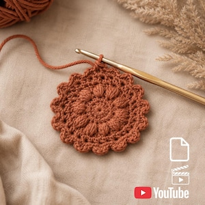May include: A circular, rust-colored crochet piece featuring a textured puff stitch pattern and a scalloped edge. A gold-toned metal crochet hook is inserted into the top of the work, with a matching yarn ball visible in the upper corner.