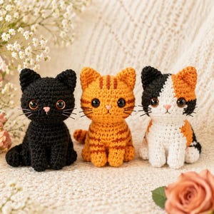 May include: Three small crocheted cat figurines sitting in a row. The first is solid black, the second is orange with darker orange stripes, and the third is a calico pattern with patches of black, white, and orange. Each has large dark eyes.