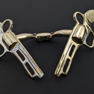 Könnte beinhalten: Zwei goldfarbene revolverförmige Objekte, die durch einen kleinen Steg verbunden sind. Die Revolver haben detaillierte Griffe und offene Rahmen. Die Objekte befinden sich auf einer schwarzen Oberfläche.