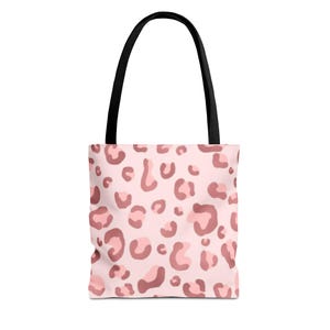 May include: A pink tote bag with a black handle. The bag features a repeating pattern of dark pink leopard spots on a light pink background. The tote bag is designed for carrying items.