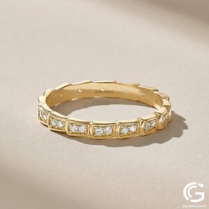 "An angled studio shot of the gold geometric diamond eternity ring, showing the pattern of diamond-paved squared links. It is set against a light beige textured background with soft lighting."