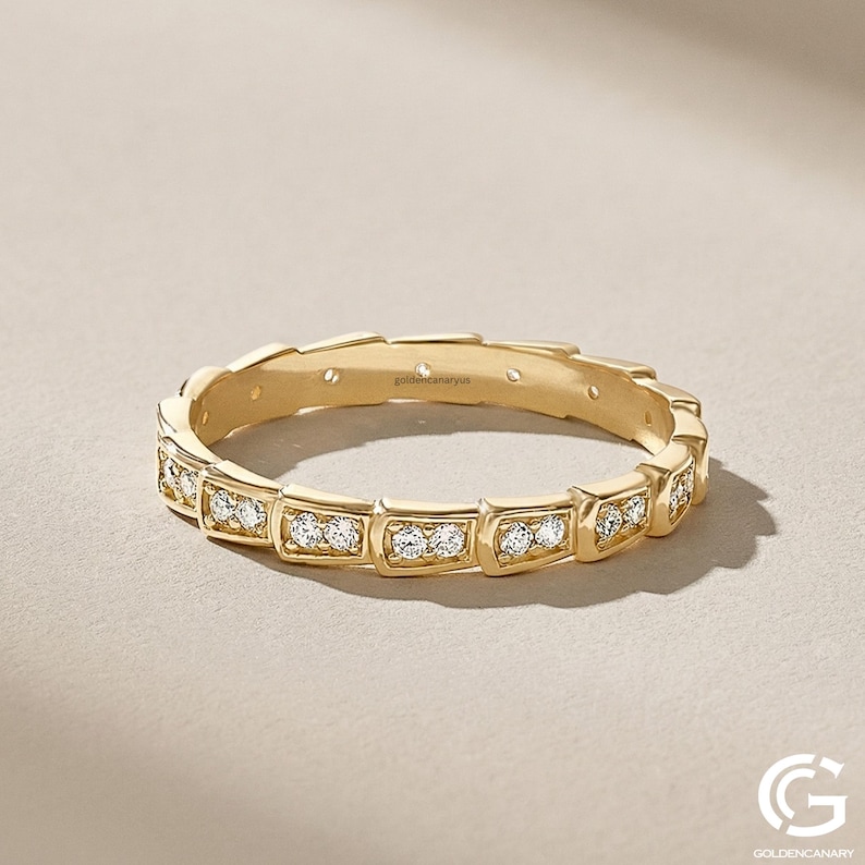 "An angled studio shot of the gold geometric diamond eternity ring, showing the pattern of diamond-paved squared links. It is set against a light beige textured background with soft lighting."