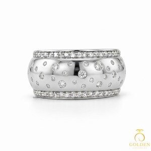 May include: A wide, silver ring with a polished finish. The ring features numerous small, sparkling diamonds scattered across the surface, with a row of smaller diamonds encircling the top and bottom edges. This ring is a piece of jewellery.