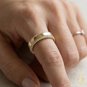May include: A gold band ring featuring a smooth, polished centre section flanked by two rows of small, clear, round-cut stones set along the edges. The ring is worn on a finger, with a second, thinner silver-toned band visible on another finger.