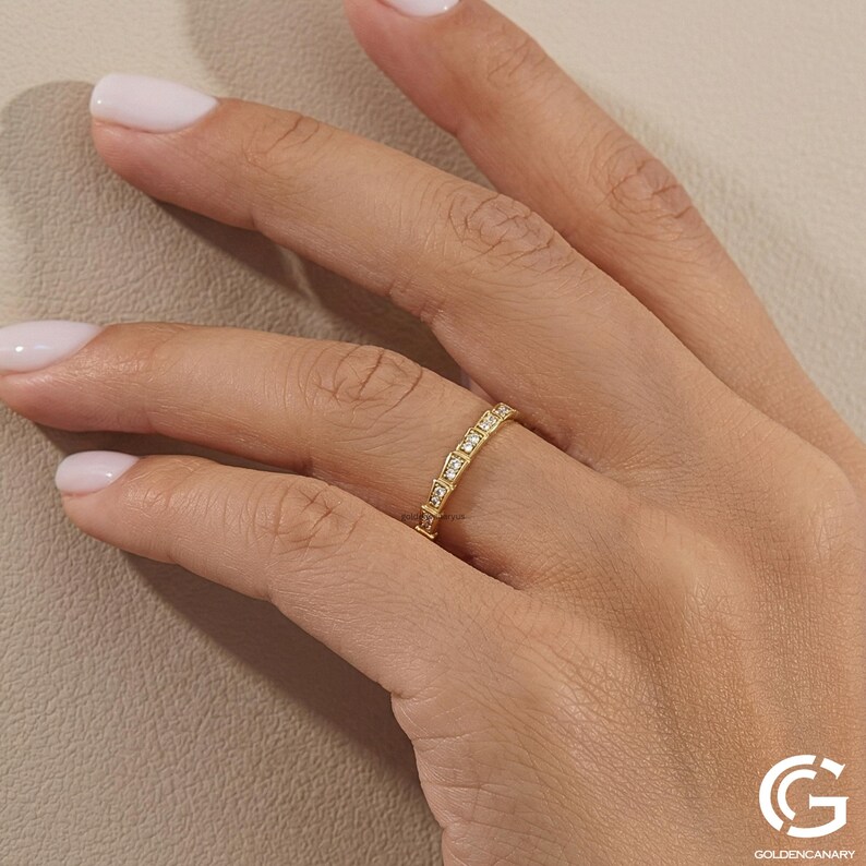 "Close-up shot of a hand with a pale-pink manicure wearing the gold diamond eternity ring, showcasing how the geometric band looks when worn on a finger. A soft shadow is cast on the textured background."