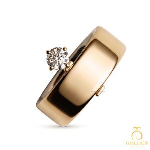 May include: A wide, polished gold band ring featuring a single round, brilliant-cut white diamond set in a four-prong setting on one side of the band. The ring has a smooth, reflective surface and a small, subtle metallic tab on the opposite side.