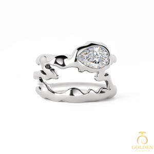 May include: A silver ring set featuring a pear-shaped diamond. The ring has a unique, organic design with a flowing, molten metal appearance. The band is split into two parts, creating a layered effect. The ring is set against a white background.