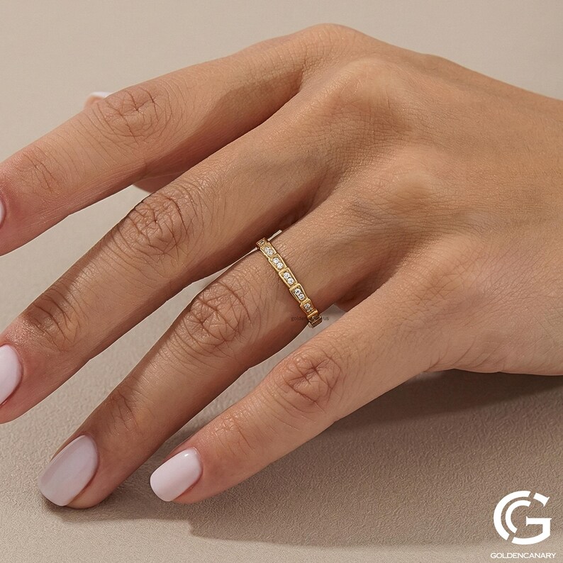 "A photograph of a hand with neat, light-pink manicured nails wearing the gold geometric diamond ring on the ring finger. The hand is posed elegantly against a beige textured backdrop. The ring's square diamond-paved links are visible."