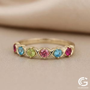 May include: A gold band ring featuring a series of colorful gemstones. The ring showcases a row of round-cut gems in shades of pink, blue, green, and red. The ring is a delicate piece of jewelry.