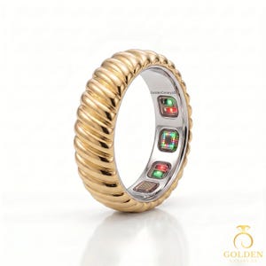 Könnte beinhalten: Ein goldfarbener Ring mit einer gerippten, strukturierten Außenfläche. Die Innenseite des Rings weist eine glatte, silberfarbene Auskleidung mit drei eingebetteten elektronischen Sensormodulen auf, die kleine rote und grüne Lichter zeigen.