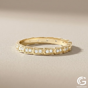 "Close-up product photograph of a 14k yellow gold eternity band. The ring features a unique geometric, squared-box segment design, with each segment paved with small, brilliant-cut diamonds. The ring is resting on a neutral, textured beige background. The goldencanaryus brand name is engraved inside the band. A 'G' logo is visible in the corner."