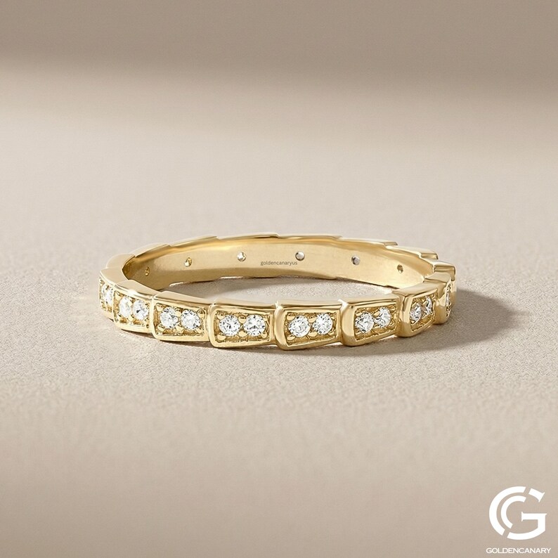 "Close-up product photograph of a 14k yellow gold eternity band. The ring features a unique geometric, squared-box segment design, with each segment paved with small, brilliant-cut diamonds. The ring is resting on a neutral, textured beige background. The goldencanaryus brand name is engraved inside the band. A 'G' logo is visible in the corner."