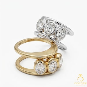 Peut inclure: Deux bagues, une en or et une en argent, chacune ornée de trois pierres précieuses ovales. La bague en or a une finition polie, tandis que la bague en argent a une surface brillante et réfléchissante. Les bagues sont sur fond blanc.