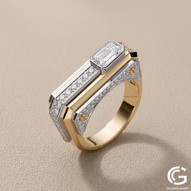 Angled view of a modern, geometric men's ring in two-tone yellow and white gold. The ring features an off-center emerald-cut diamond, a row of pave-set diamonds along the top, and industrial-style gold screw details on the side panels.