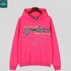 May include: A bright pink hoodie with a front pocket and a hood. The hoodie features the word "sp5der" in a stylized font, along with a spiderweb design and text that reads "defended the punk" and "bares the alibi".